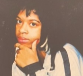 Chanell has a serious look on her face. Her chin is resting on her hand. She is wearing a white shirt with a stripe down the arm.